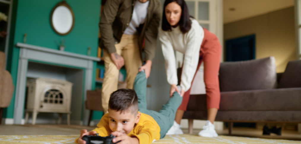 Boy lying face down on the floor while holding a gaming console and mom and dad pulling his leg