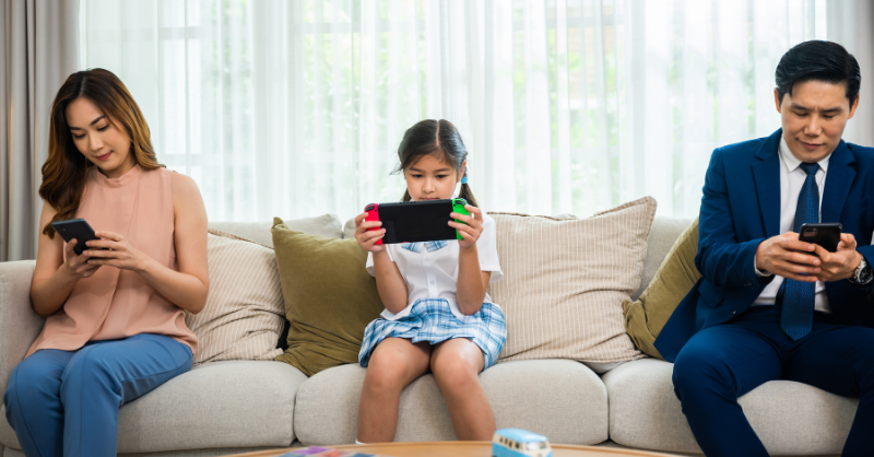 Girl sitting in a couch and in between parents. All of them seems occupied with their gadgets