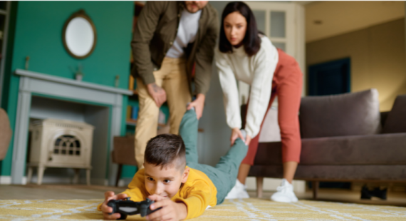 Boy lying face down on the floor while holding a gaming console and mom and dad pulling his leg