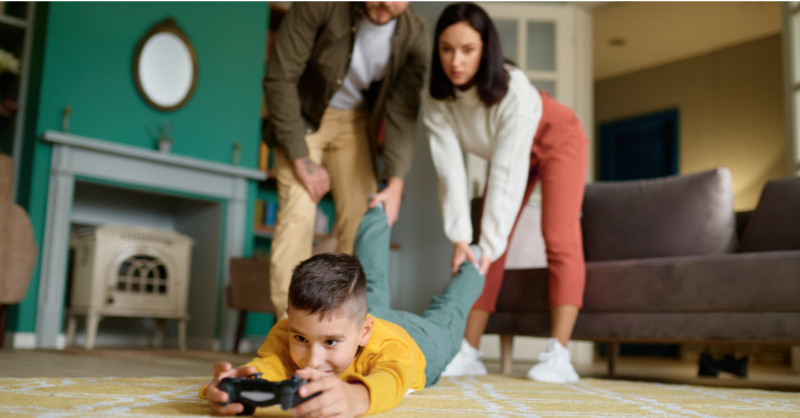 Boy lying face down on the floor while holding a gaming console and mom and dad pulling his leg